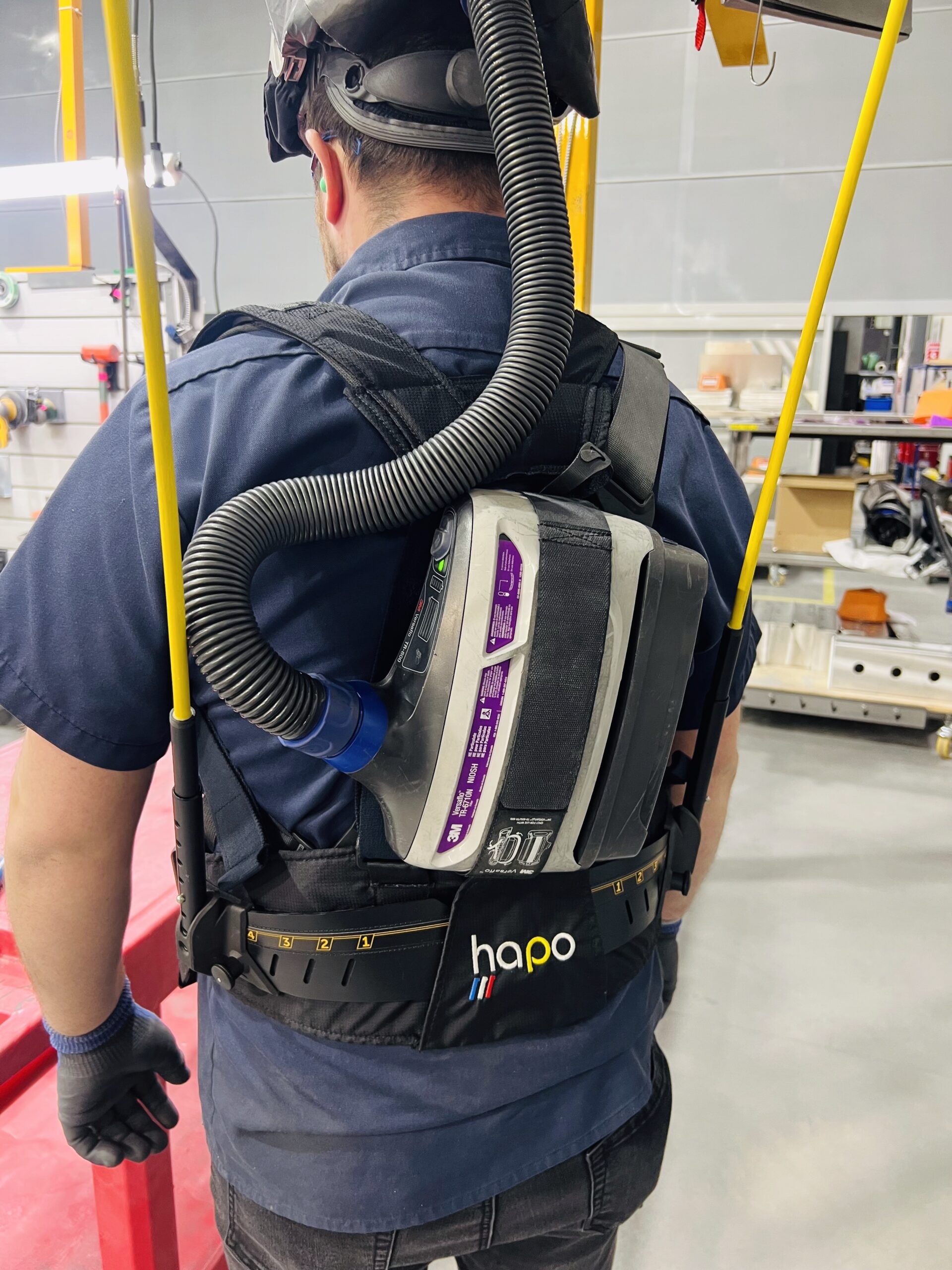 Rear view of an operator wearing both a Hapo Front exoskeleton and a 3M Versaflo powered respirator — PPE compatibility demonstrated